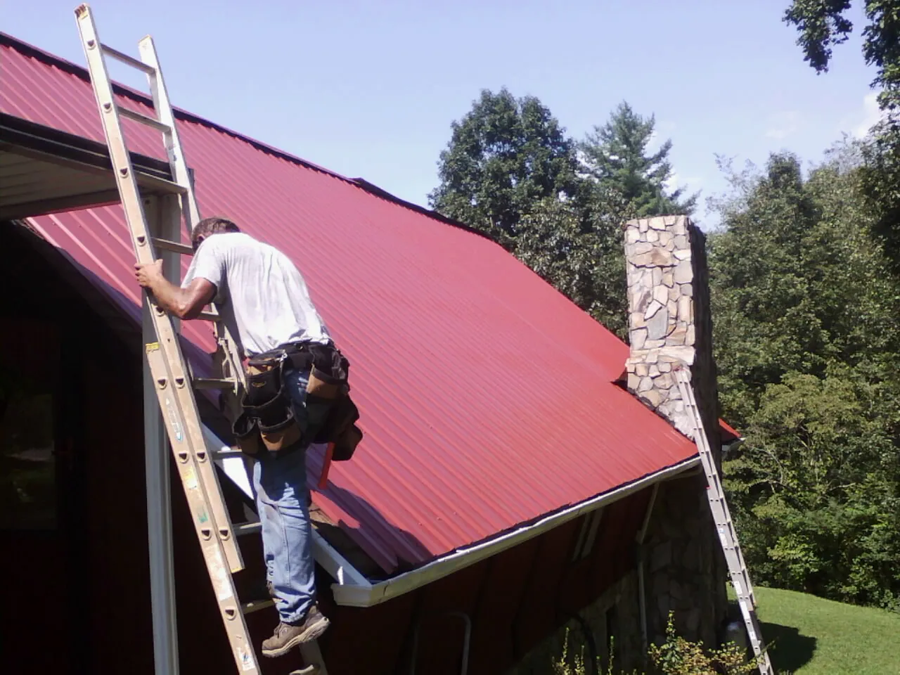 Roofer on a ladder beside a red metal roof and stone chimney — Soffit & Fascia Repair by Roofing Brothers in Descanso